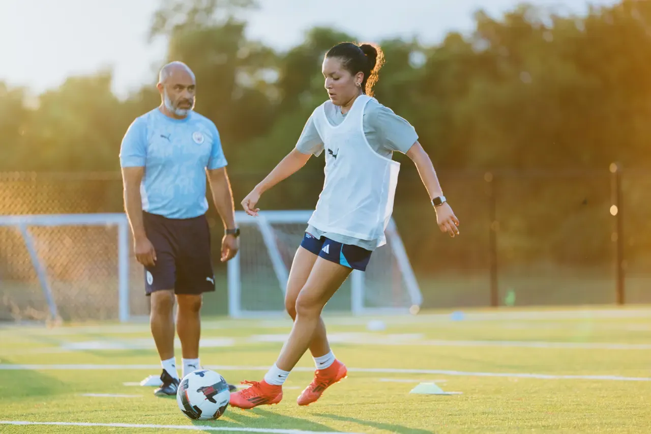 Coach Alberto Carbajal observing a player working with the ball on the training pitch at sunset