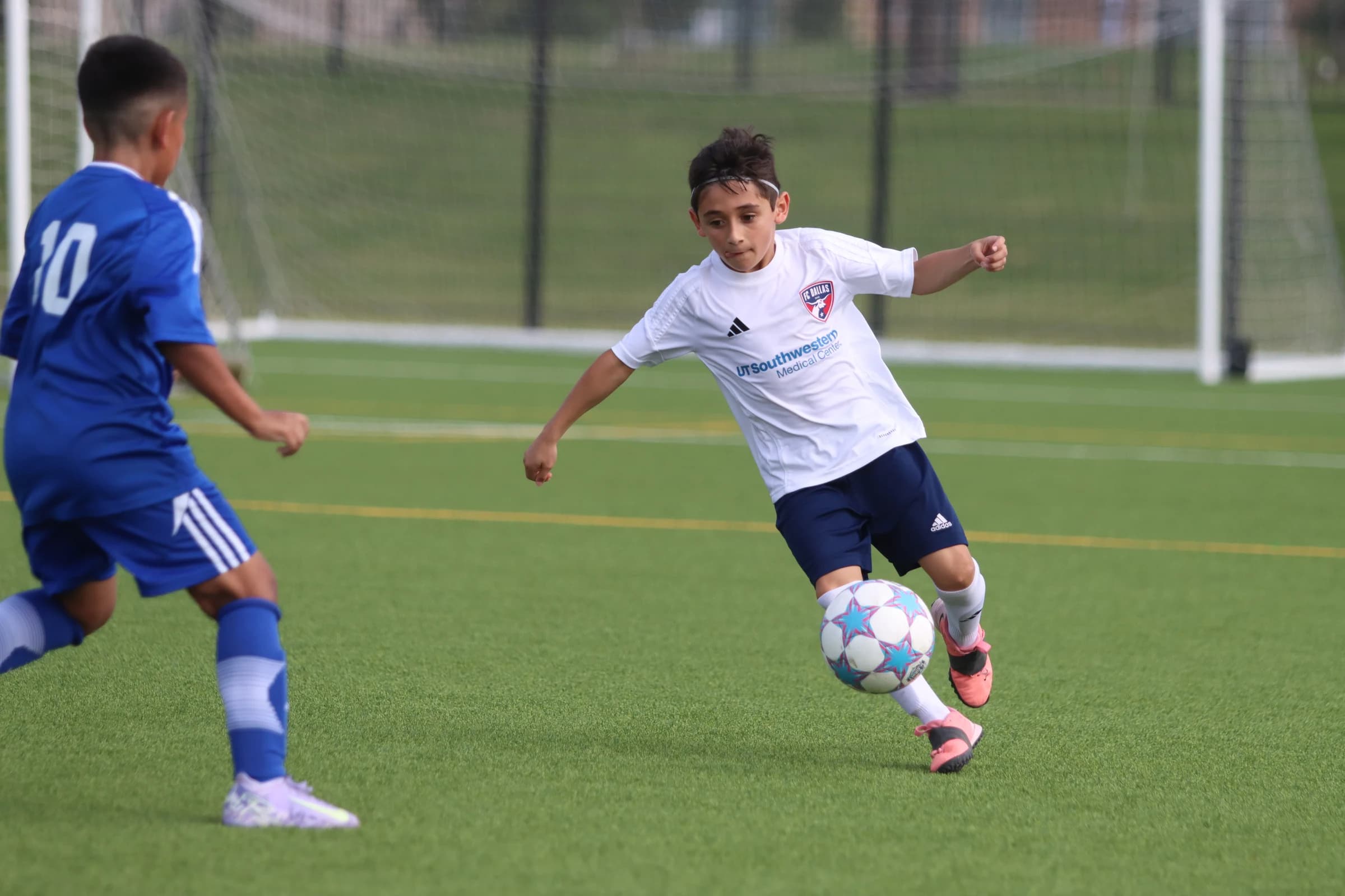 A youth footballer in a white FC Dallas shirt controlling the ball while facing an opponent in blue
